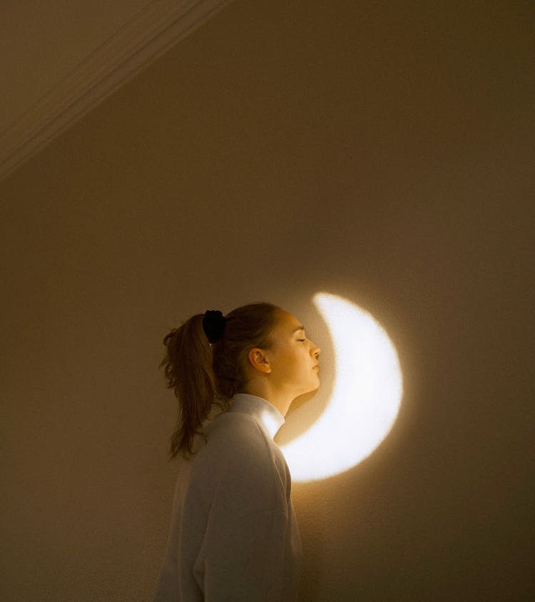 Portrait of a caucasian girl with brunette hair leaned against a wall. Her eyes are closed and there is the shape of a moon projected onto the wall. It looks like she is resting her head on the moon. The tones of the image are warm and yellow.