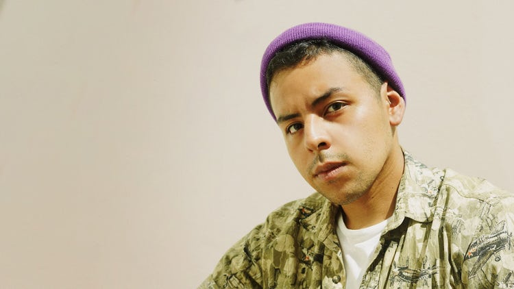 Young Latin Peruvian, dark with tattoos, in a green shirt, white polo shirt and purple beanie.  He looks short hair and a direct look at the camera.