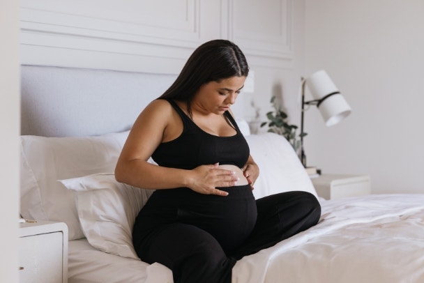 Pregnant Carribbean Indigenous woman practicing self care and putting lotion on baby bump in bed