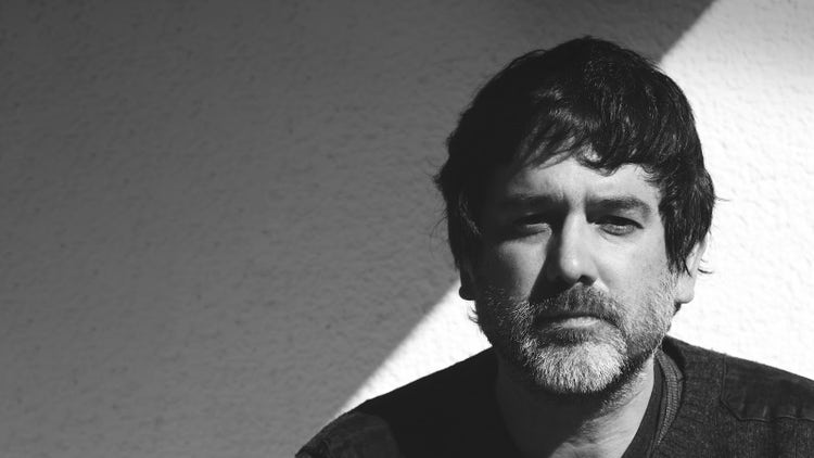 Photograph of Carlos David. Close up, horizontal black and white headshot. A serious man with a short beard and short textured fringe looking at the camera. He is wearing a dark-gray lambswool sweater on top of layered clothing against a white background diagonally divided in light and shadow—hard sunlight.