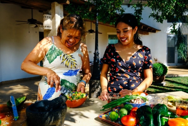 Happy young woman standing by mother preparing Mexican salsa at kitchen counter in backyard on sunny weekend