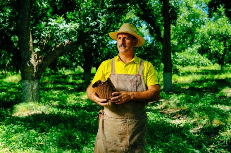 Mature male farmer wearing apron and hat looking away while holding rusty metallic can against trees at orchard