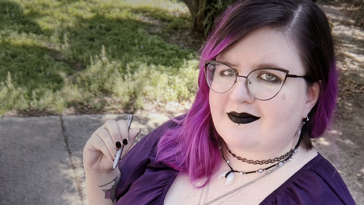 A selfie photo of Michaela, a fat white woman with black hair that fades to vibrant purple. She is wearing glasses, multiple necklaces, and has subtle face piercings. She is wearing black lipstick that matches her nail polish, a purple shirt, and she is holding a pen in her right hand. She is sitting in her wheelchair outside.