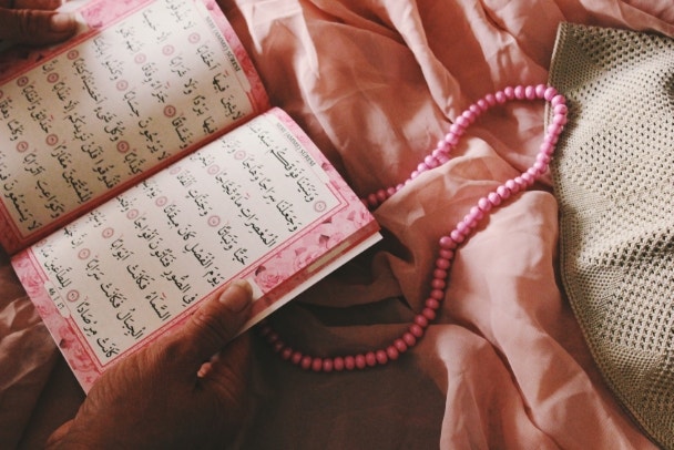 womans hands holding a copy of Quran, book of Islam religion, papers with prayers in Arabic language, with pink rosary and taqiyah in pink textile background