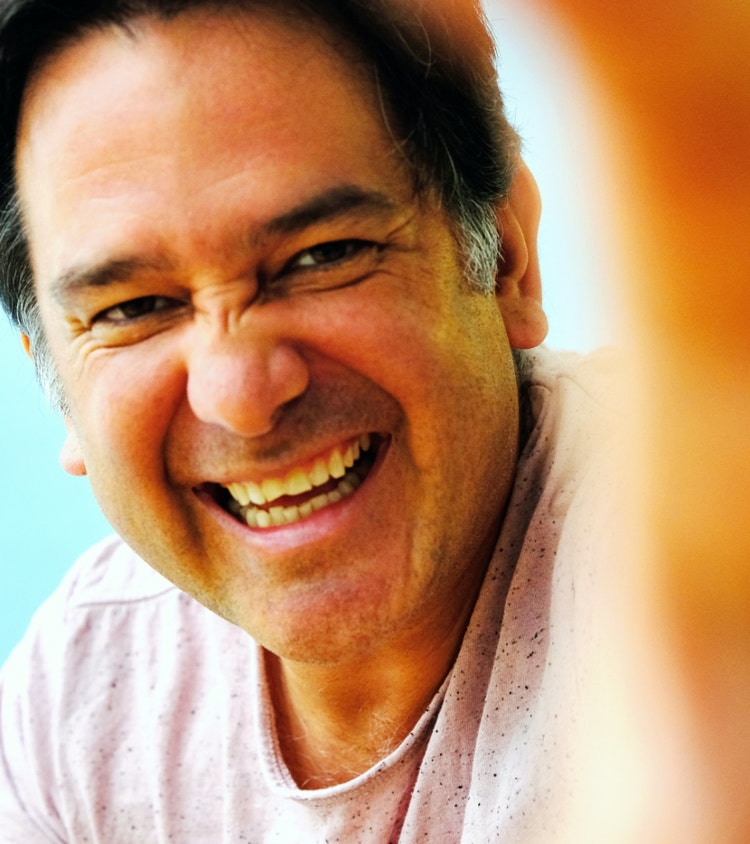 Photograph of Carlos David. Close up, vertical color headshot. Smiling adult man. No beard and black hair with salt and pepper short sideburns. Wearing a pale pink t-shirt with tinny black dots. His blurred palm is covering one quart of the right side of the frame. 