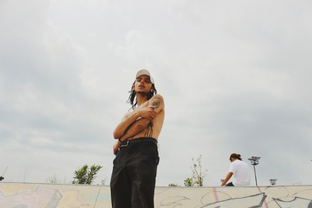 young man in skatepark, shirtless with tight hair braids and tattoos with friend in urban background