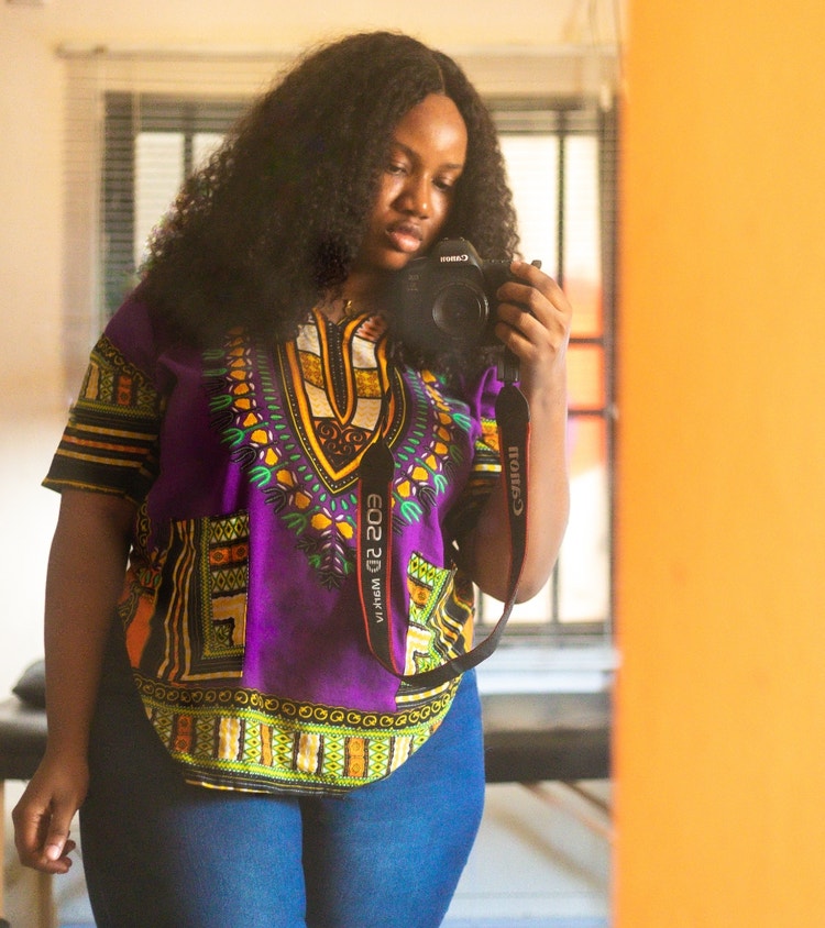 Black African woman with a curly wig on holding a camera, taking a mirror selfie with her canon camera. She is wearing a purple patterned “dashiki” and dark blue pair of jeans. Holds her camera with her right hand, while her other hand is on her side. (A dashiki is a colorful garment worn mostly in West Africa)