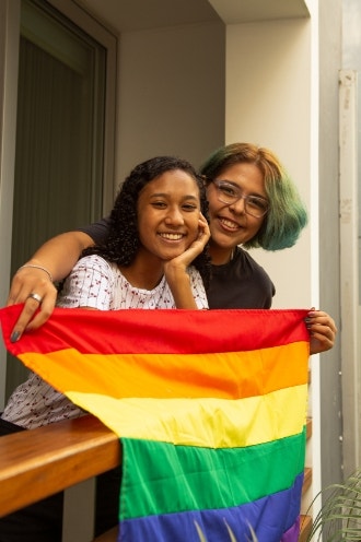 Pareja de lesbianas jóvenes abrazadas sosteniendo la bandera LGTBQ en su terraza