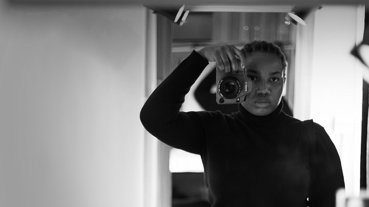 Black African woman with cornrows wearing a black turtle neck knitted top. She has a pair of dark blue jeans on. She is holding a canon camera & taking a mirror selfie with her camera. She is also carrying a back tote back on her left arm. Holds her camera that has a white case with her right hand, while her other hand is on her side.