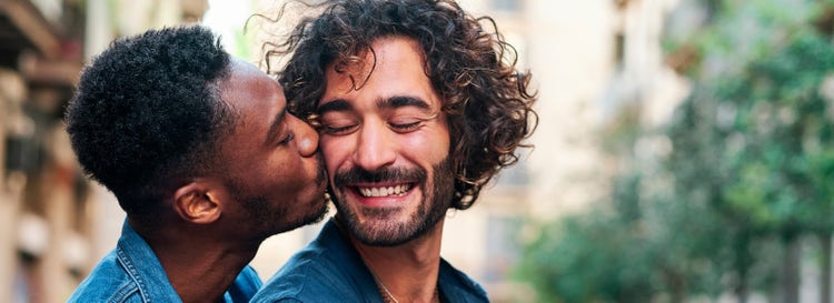 Closeup portrait of gay men in denim. One smiles as boyfriend kisses his cheek. Blurred street backdrop outdoors with trees.