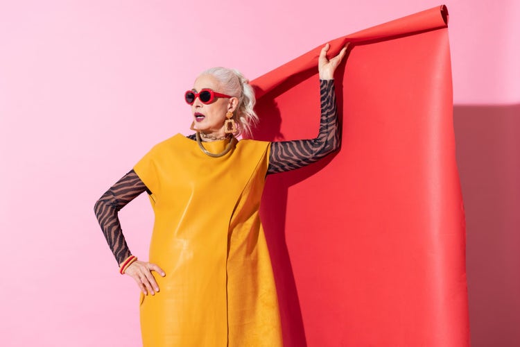 Blonde woman in eclectic yellow outfit and accessories holds red roll of studio backdrop in front of pink background.
