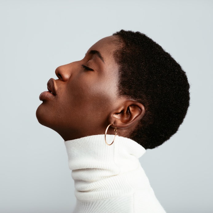 Profile portrait of Black woman with short hair, eyes closed, in white turtleneck and small gold hoop earring. Neutral studio backdrop.