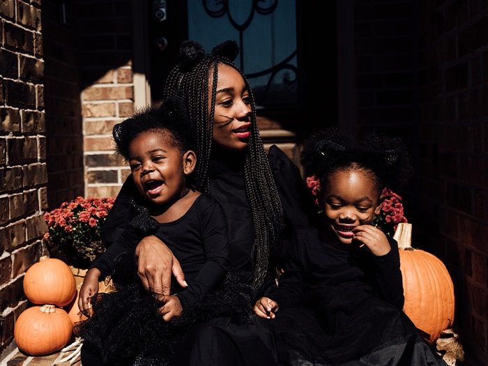 Young Black mother with two toddler daughters in cat costumes, on brick front porch area with pumpkins, chrysanthemums.
