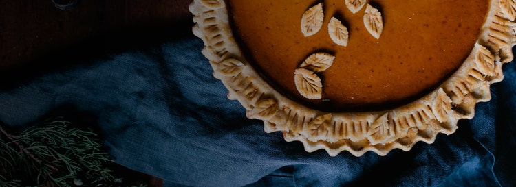 Closeup shot of pumpkin pie with leaf detailing on shell edges. Shot from above on navy fabric; rosemary bunches to side.