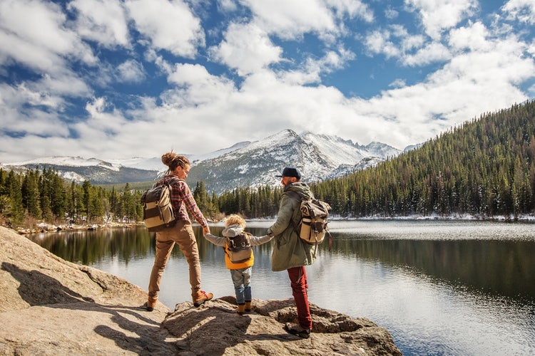 Parents each hold toddler son's hand, walking along lake in mountains, all in backpacks. Evergreen trees and sunny cloudy blue sky.