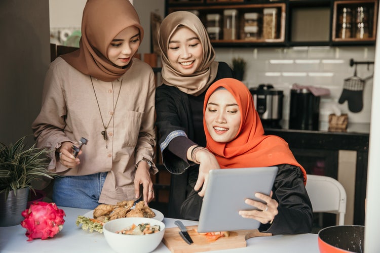 Three young women in hijab and casual clothes look at recipe on tablet while preparing meal in kitchen at table.