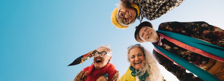 Four elderly people in semi-circle wearing eclectic accessories and pattened clothes look down at camera, smiling and laughing. Sunny blue sky.